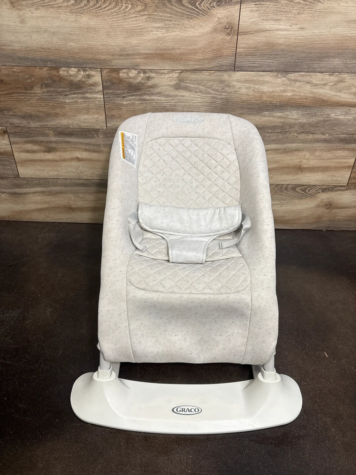 Front view of Graco JumpStart 4-in-1 bouncer in Wren showing quilted light gray seat and harness against wood wall