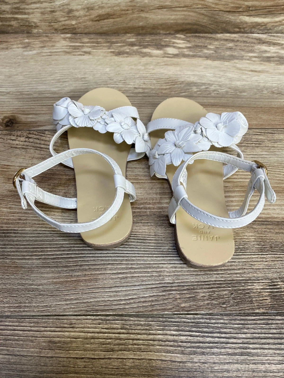 Three-quarter view of Janie and Jack 12c sandals showcasing white toe strap with flower appliques and ankle buckle on wood plank floor