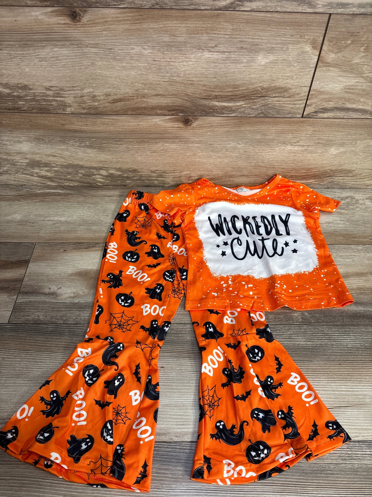 Orange Halloween kids pajamas set with ghosts, pumpkins, 'Wickedly Cute' text, and 'BOO!' print on wood floor