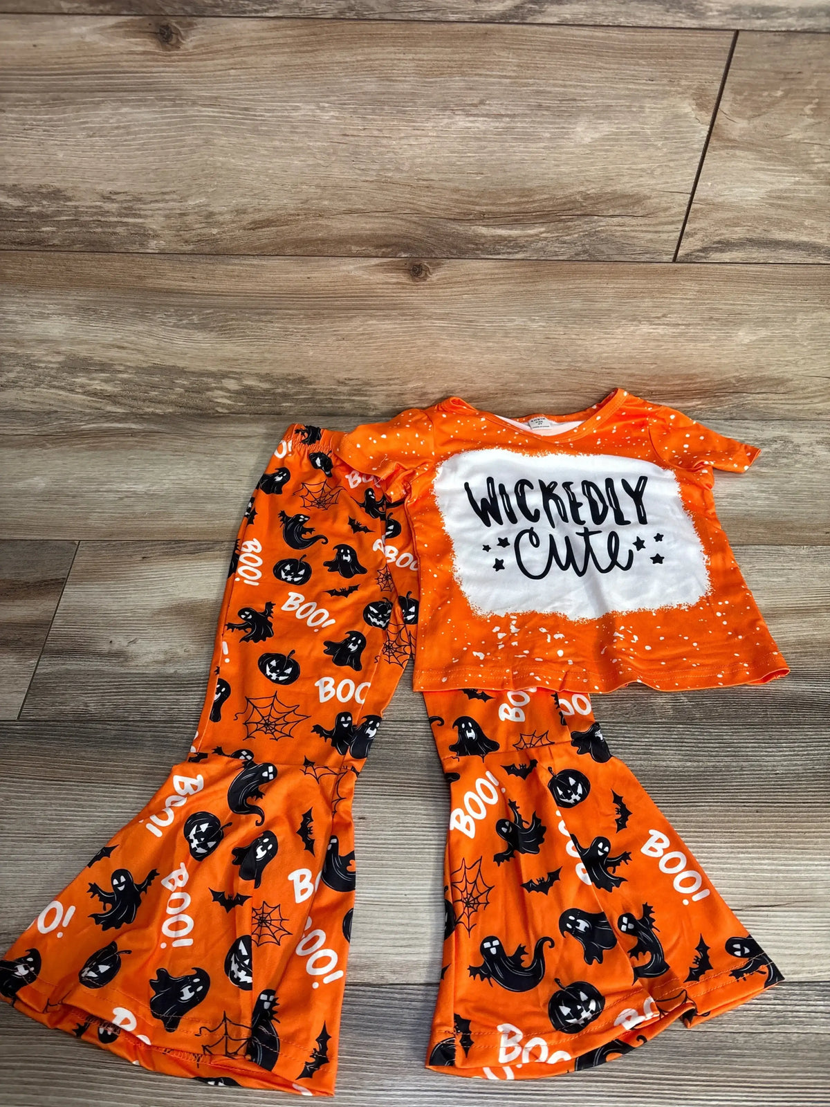 Orange Halloween kids pajamas set with ghosts, pumpkins, 'Wickedly Cute' text, and 'BOO!' print on wood floor