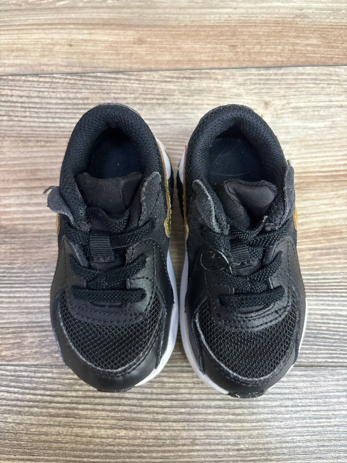 Top view of Nike Air Max Excee TD toddler sneakers, size 5C, black with gold star swoosh and white soles on wooden floor