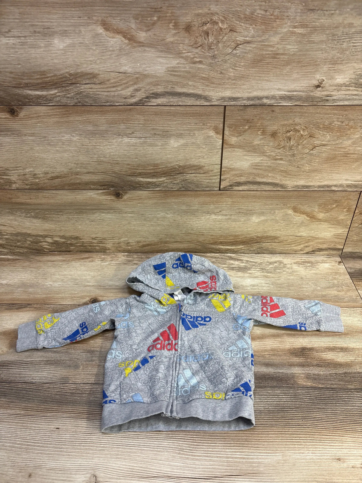 Flat lay front view of grey Adidas zip hoodie showcasing multicolor logo print with wood floor background, size 9 months