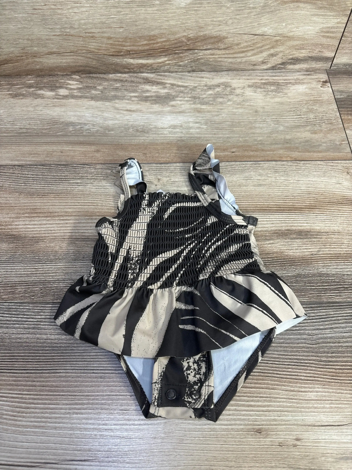 Flat lay of Carters 1pc swimsuit in brown and white zebra print showcasing smocked bodice ruffle skirt and tie shoulder straps on wood plank floor