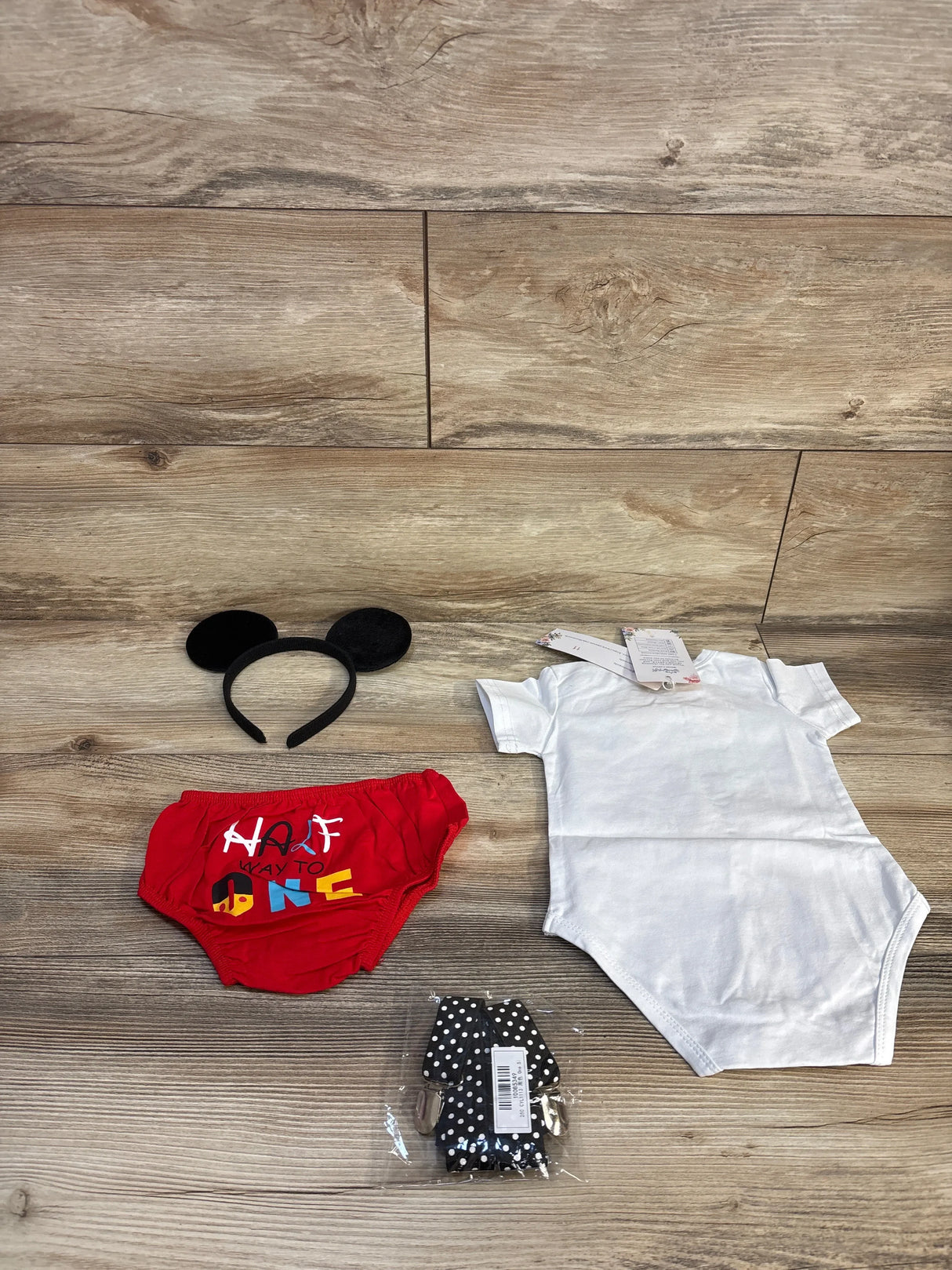 Flat-lay of 4pc suspender set: white onesie, red bloomers reading Half Way To One, black mouse ears headband on wood floor