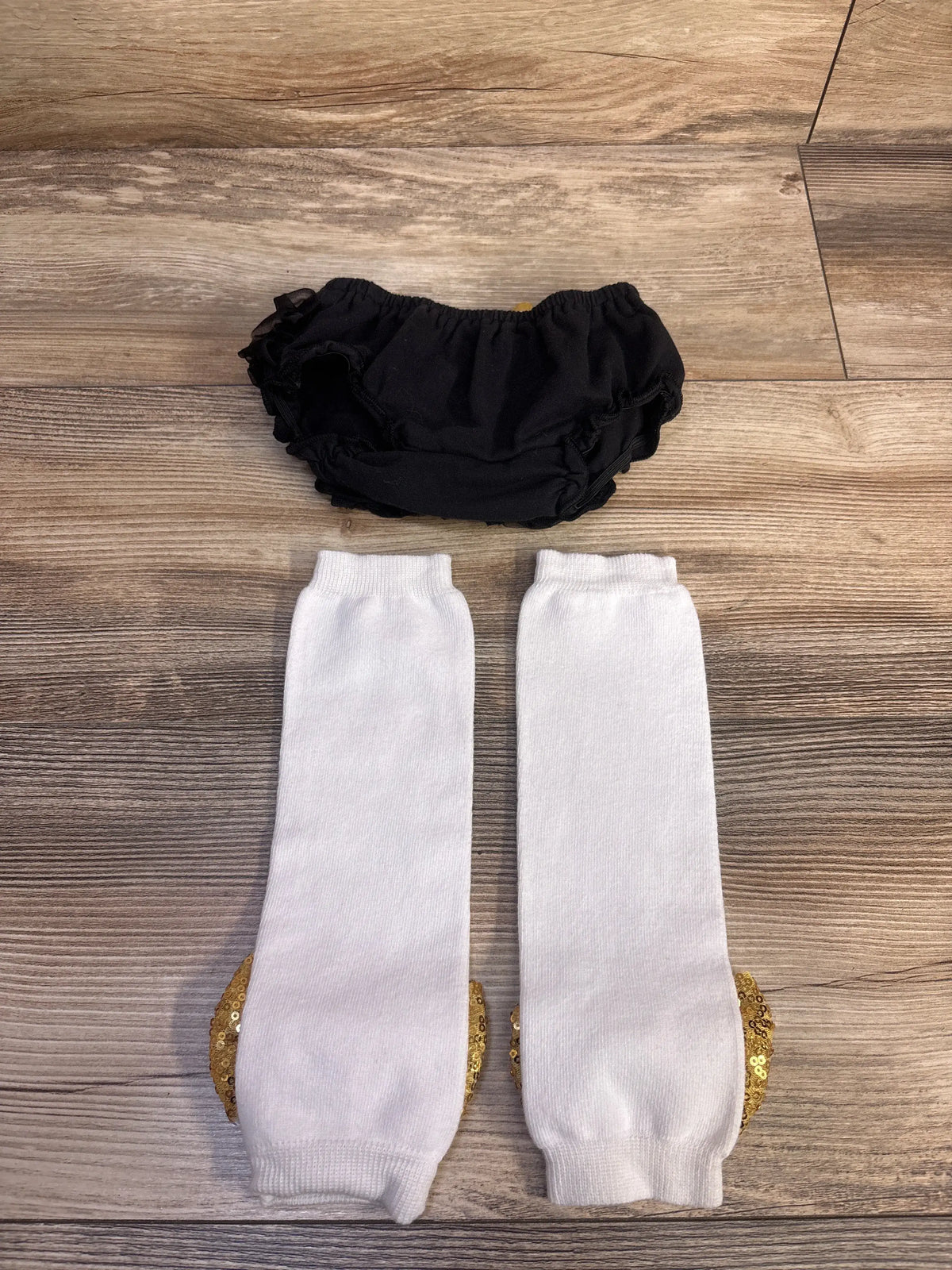 Flat lay of 2-piece black ruffle bloomers and white leg warmers showcasing ruffled edges and gold bow detailing on wood floor