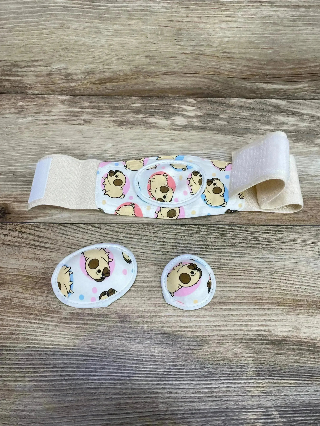Top view of Bojje umbilical hernia belt with beige Velcro strap, two padded inserts, and puppy print fabric on wood background