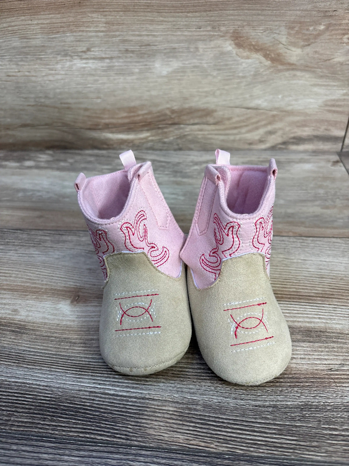 Front view of Stepping Stones cowgirl crib boots in pink and cream showcasing western embroidered designs on a wood grain surface