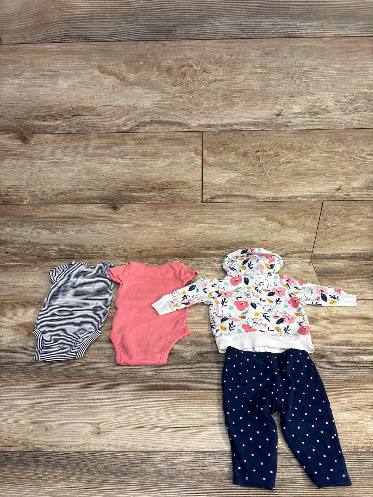 Flat lay of Simple Joys 4-piece baby set: two bodysuits, floral hooded full-zip jacket, and navy polka-dot pants on wood floor