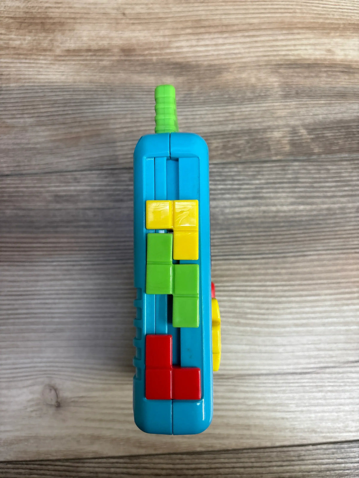 Side view of Fisher Price Laugh and Learn Lil Gamer toy in blue with yellow green and red block buttons and green antenna on wood plank floor