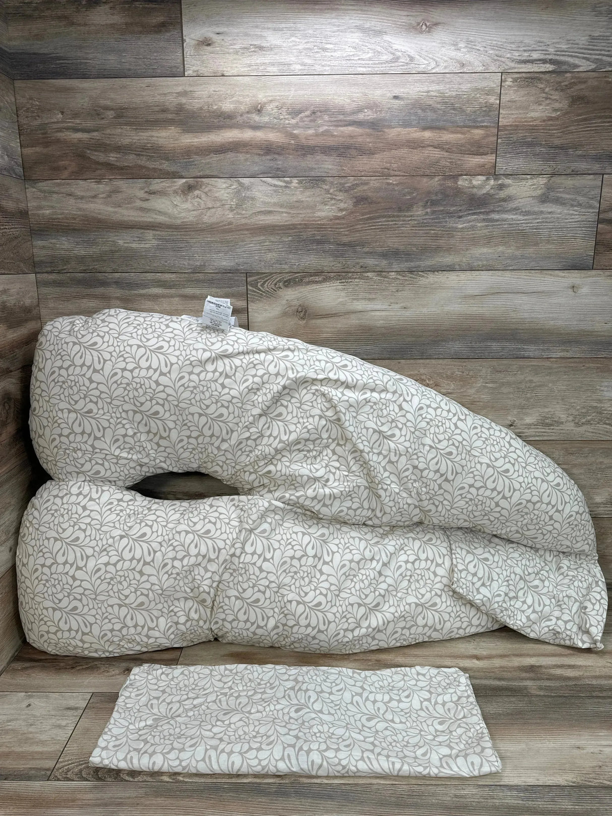 Angled view of Leachco Back 'N Belly maternity pillow showing U-shape and taupe Chic Splash pattern with matching pad on wood-look floor