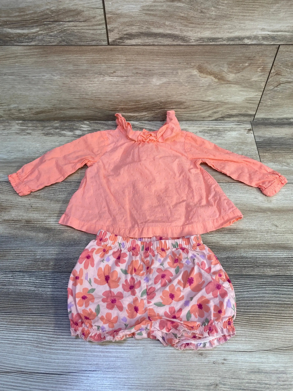 Flat lay of coral baby 2-piece: ruffled swiss-dot long-sleeve top and floral elastic shorts on wood background, 6M