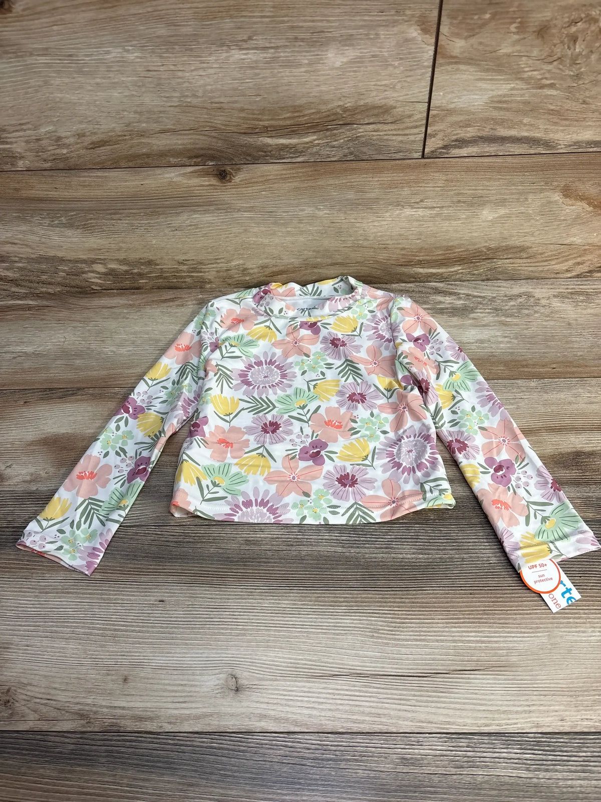 Front view of toddler long-sleeve floral rashguard showing pastel flower print and UPF 50+ tag on wood floor background