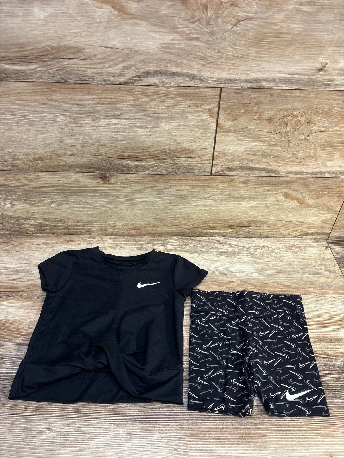 Flat lay of Nike Dri-Fit black tee and matching black shorts with white swoosh logos and repeated swoosh print on wood floor