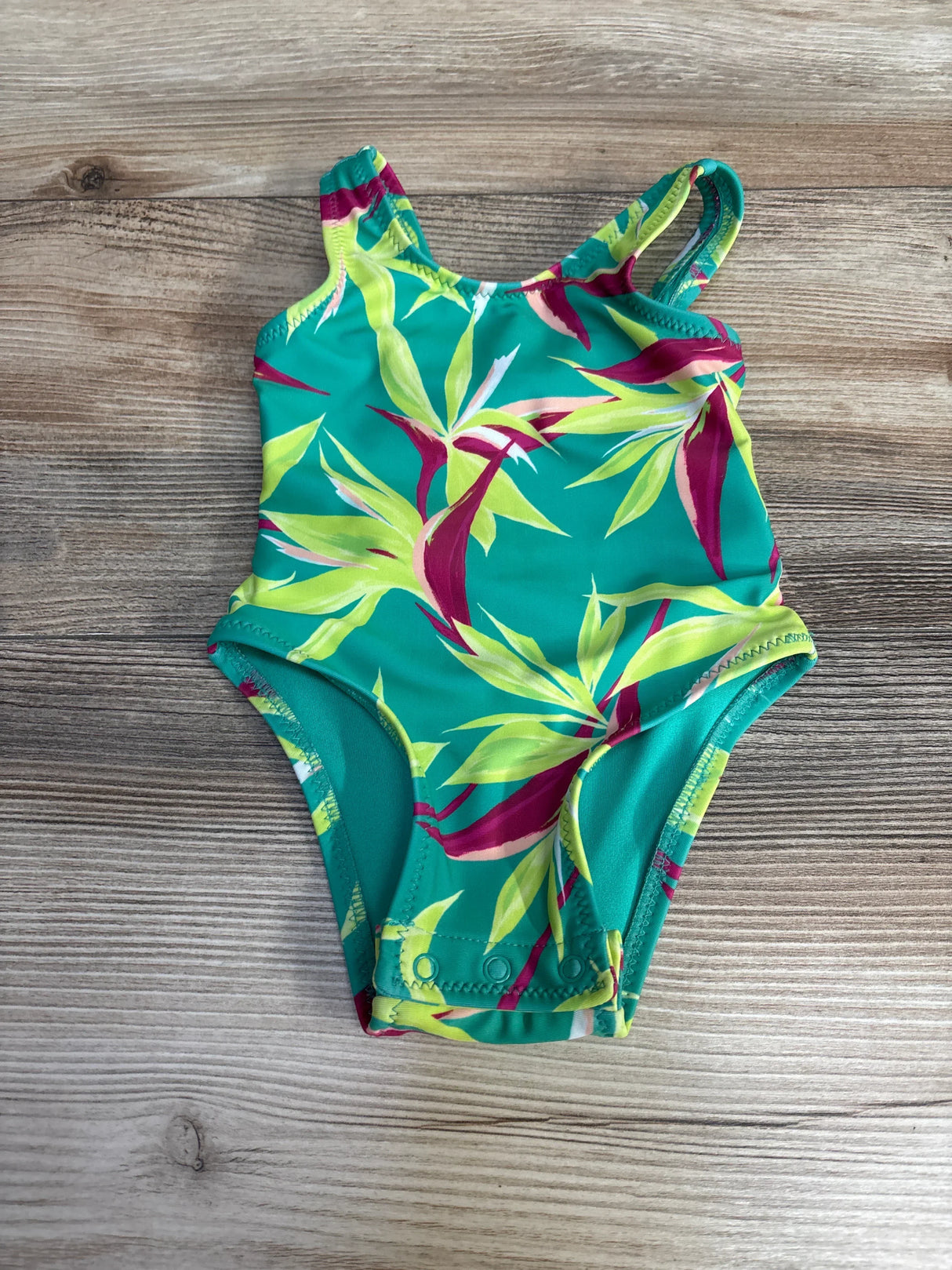 Top-down view of a green one-piece baby swimsuit showing tropical leaf print and snap crotch closure on a light wood background