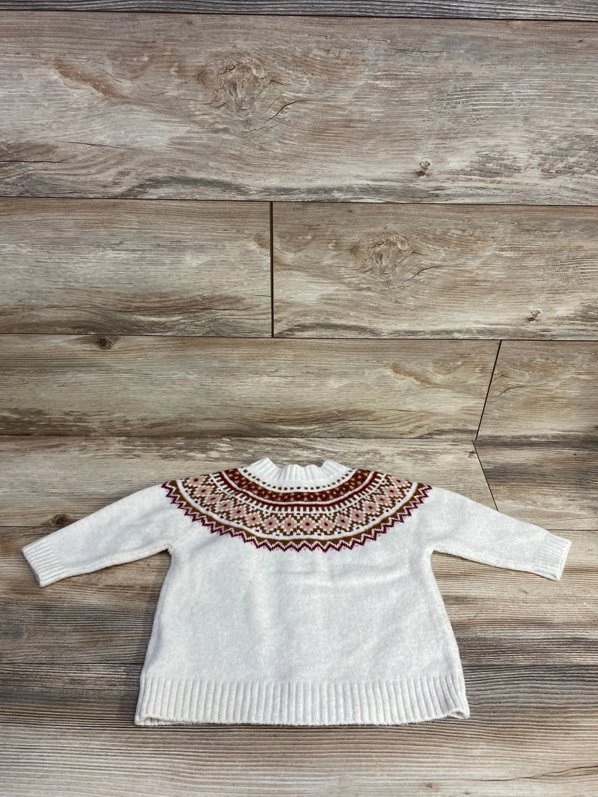 White knit baby sweater with colorful geometric yoke pattern, long sleeves, laid on wood floor