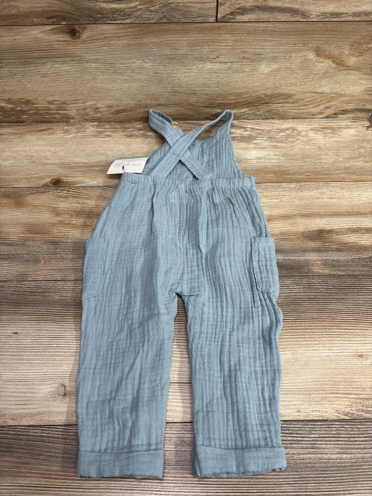 Front view of blue Little Planet organic cotton gauze henley overalls with crossed straps and side pockets on wood grain