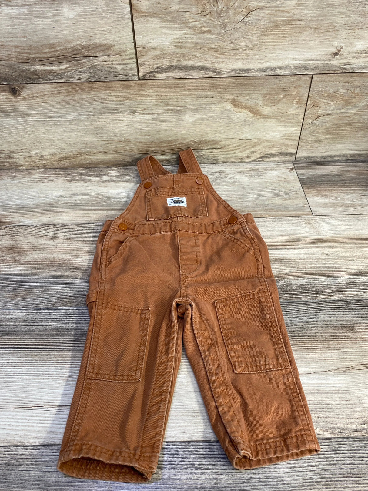 Flat-lay of brown Old Navy twill toddler overalls size 12-18 months showing bib pocket, knee patch pockets and button shoulder straps on wood background