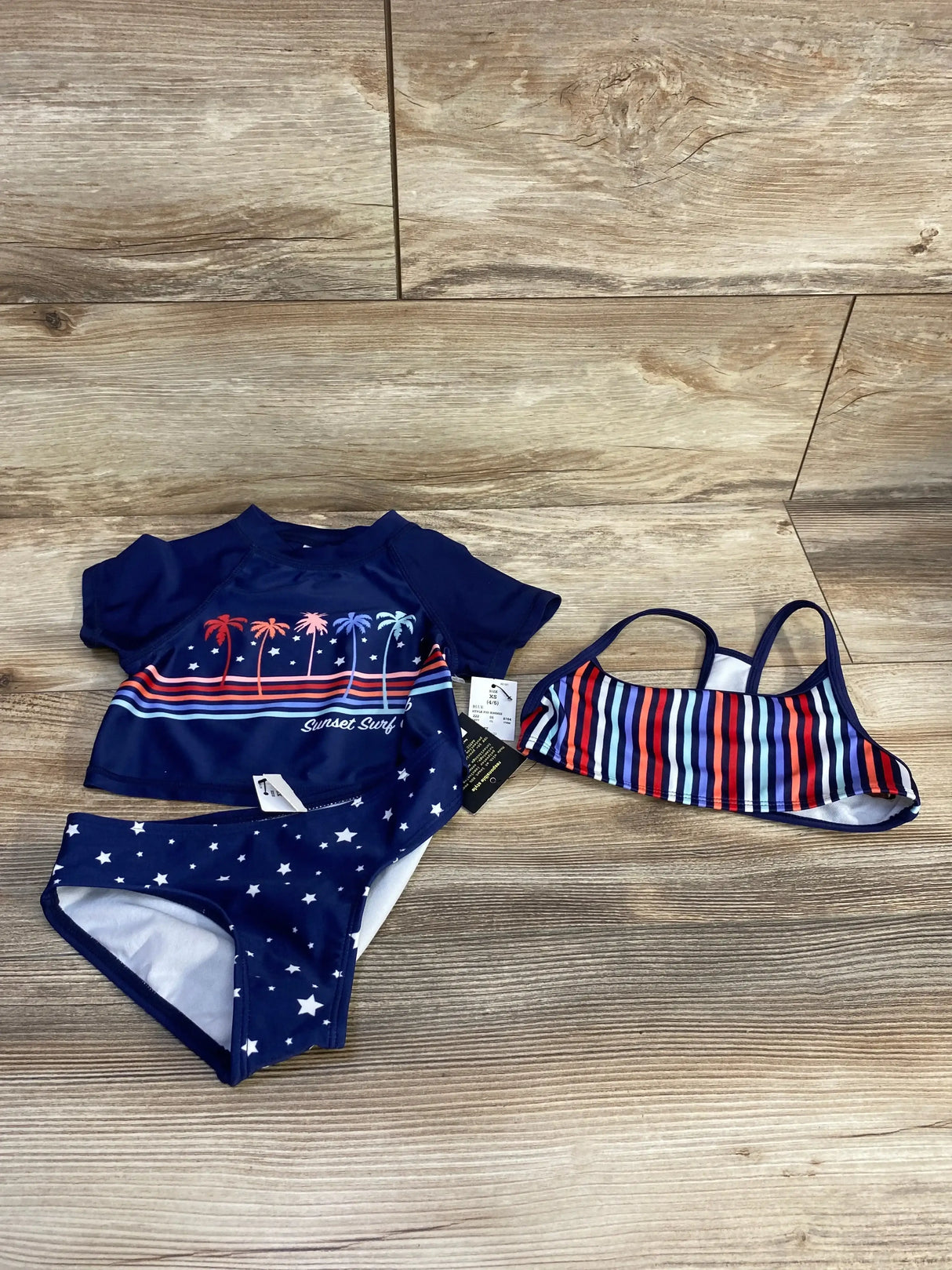 Flat lay of navy 3-piece rashguard set with palm tree Sunset Surf Club top, star-print bottoms and striped top on wood