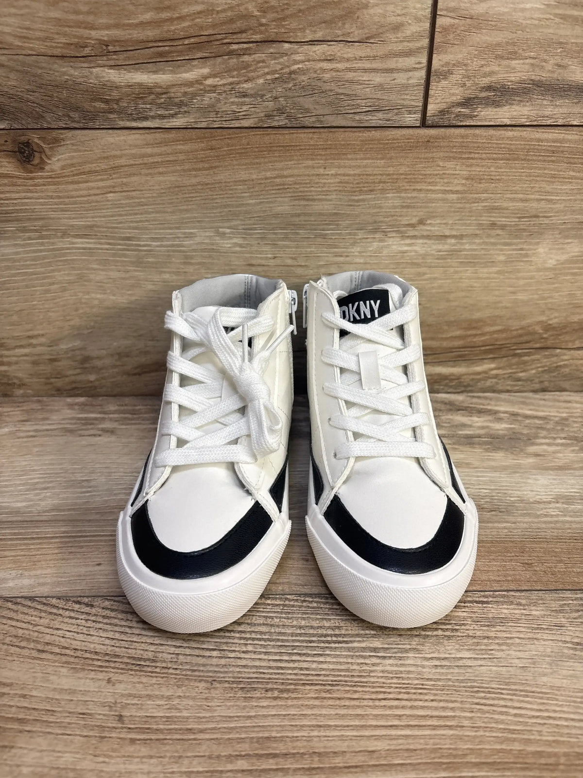 Front view of DKNY girls' Hannah Leena white high-top sneakers with black toe trim and side zipper on wood floor