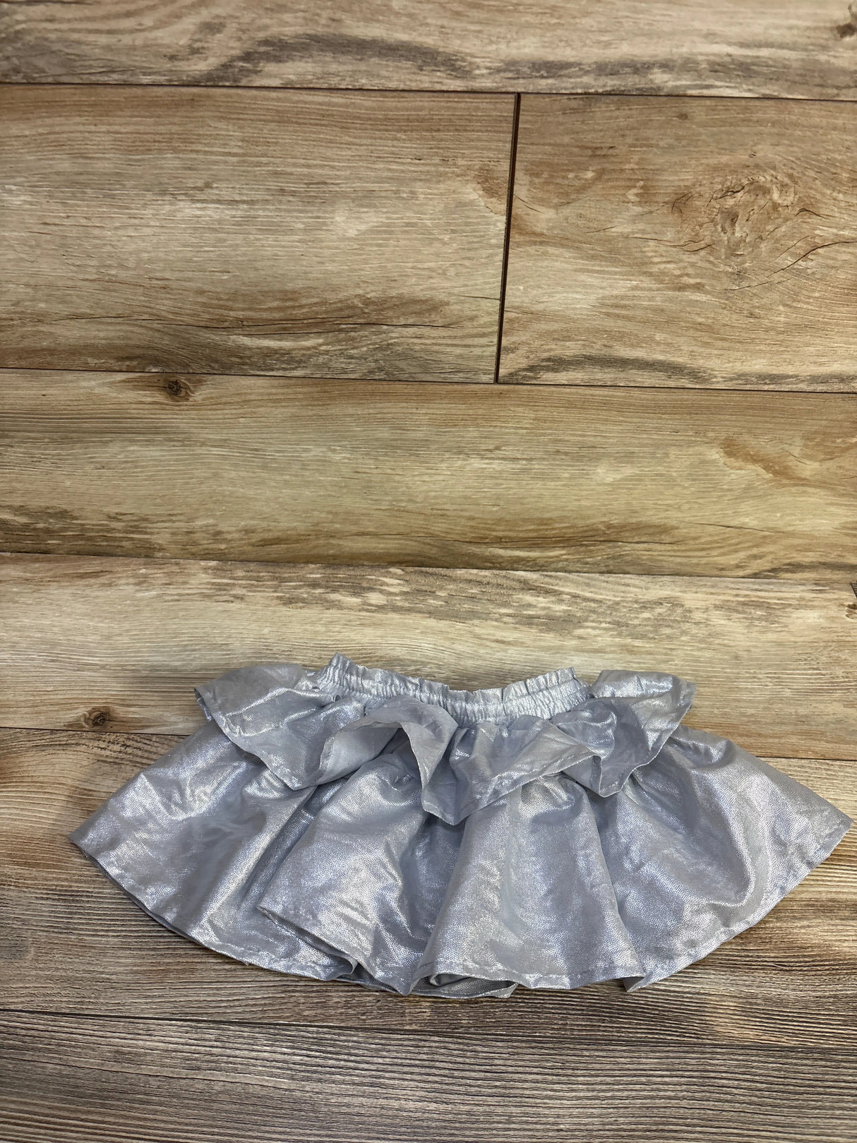 Top-down view of silver metallic toddler skort showing ruffled overlay and elastic waistband on wood floor background