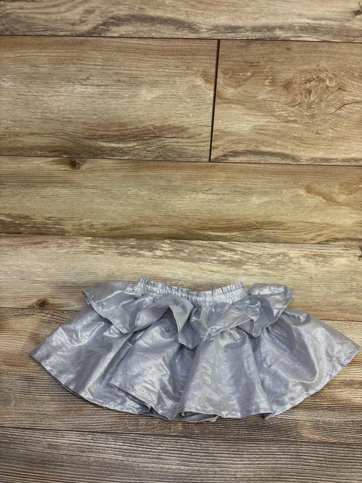 Top-down view of silver metallic toddler skort showing ruffled overlay and elastic waistband on wood floor background