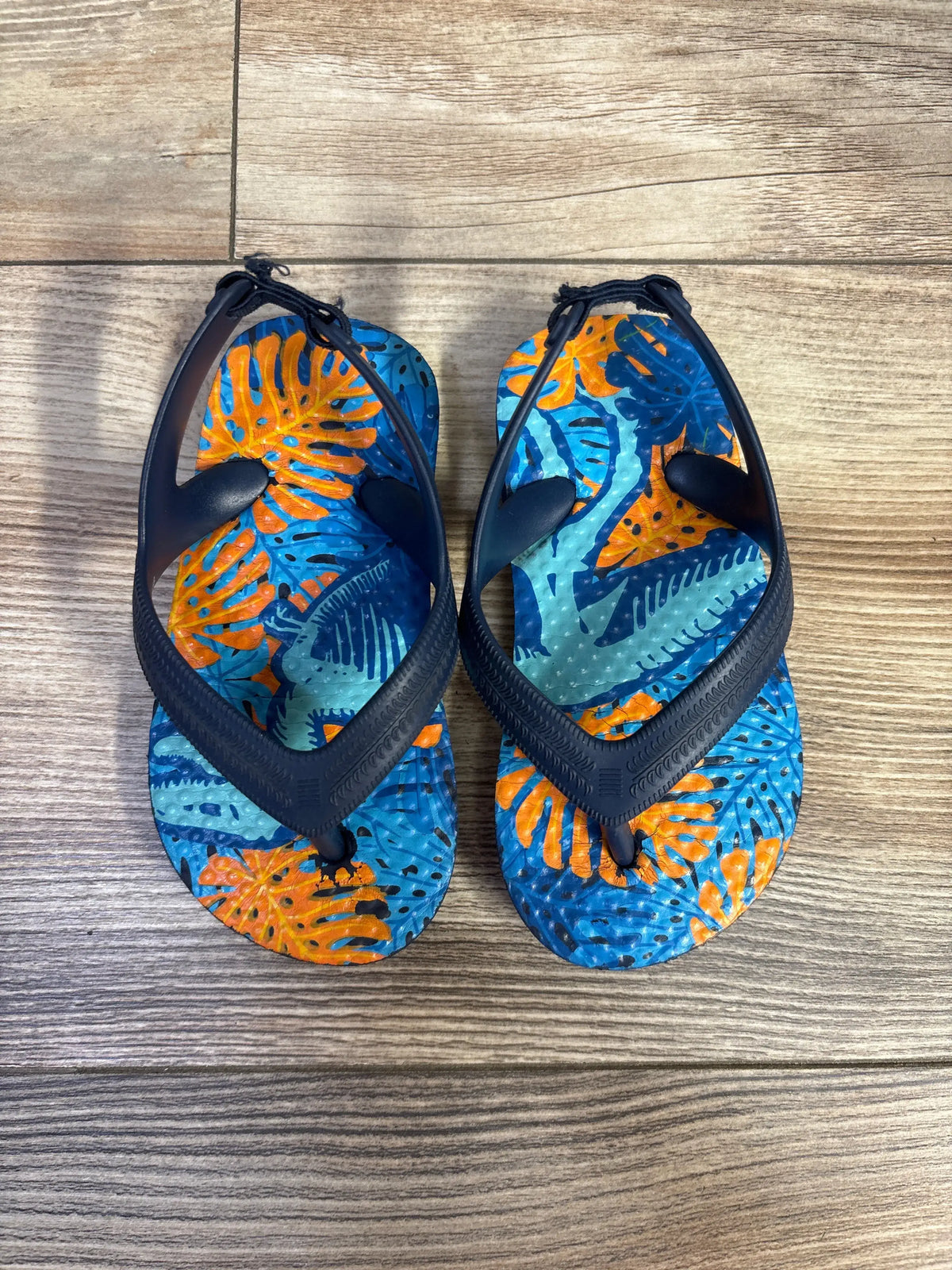 Top-down view of Wonder Nation flip flops showing navy straps and blue tropical leaf footbeds with orange accents on wood floor