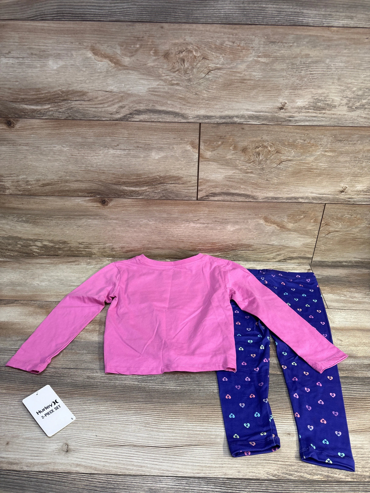 Flat lay of Hurley 2-piece long-sleeve shirt and leggings set showing pink shirt and navy leggings with multicolored heart print on wood background
