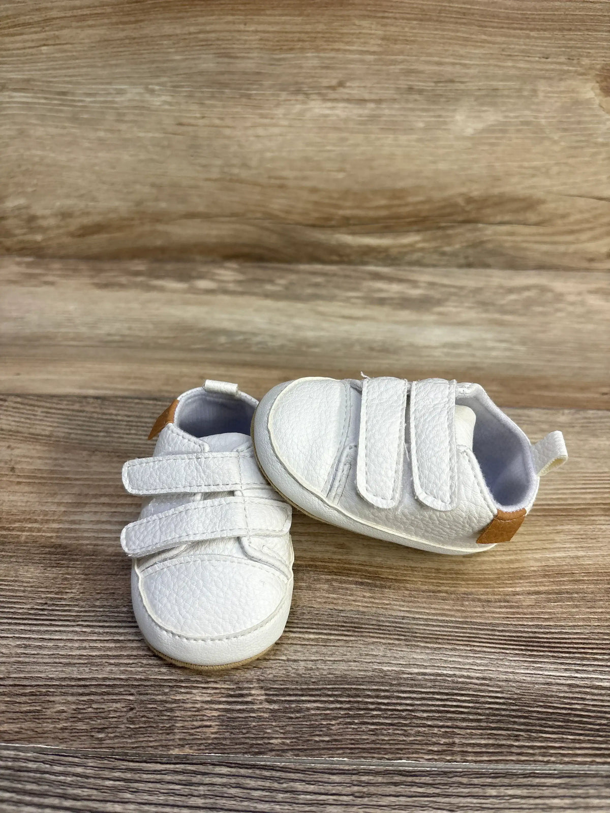 Front view of Myggpp First Walker baby sneakers in white showing two Velcro straps, pebbled faux-leather texture and tan heel tab on wood-grain background
