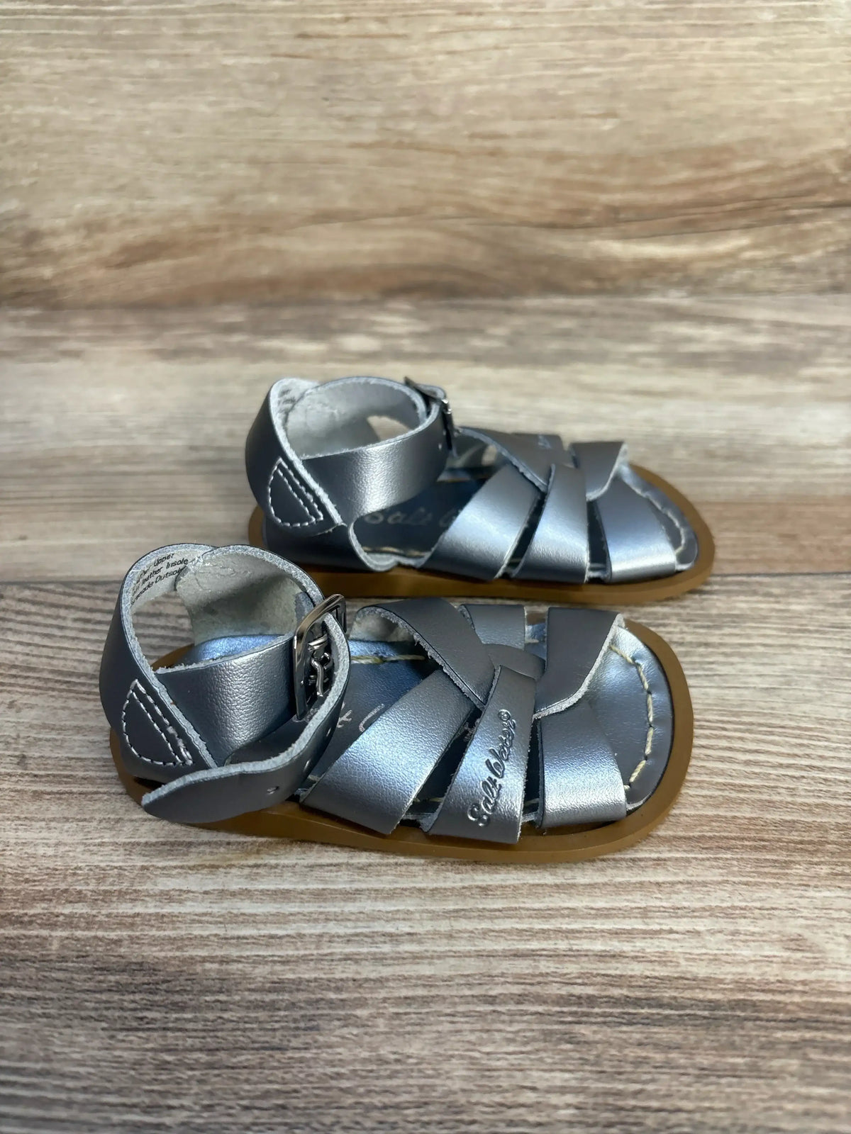 Top-down view of Salt-Water Original sandals in silver showing crisscross leather straps, buckled ankle strap, and brown sole on wood background