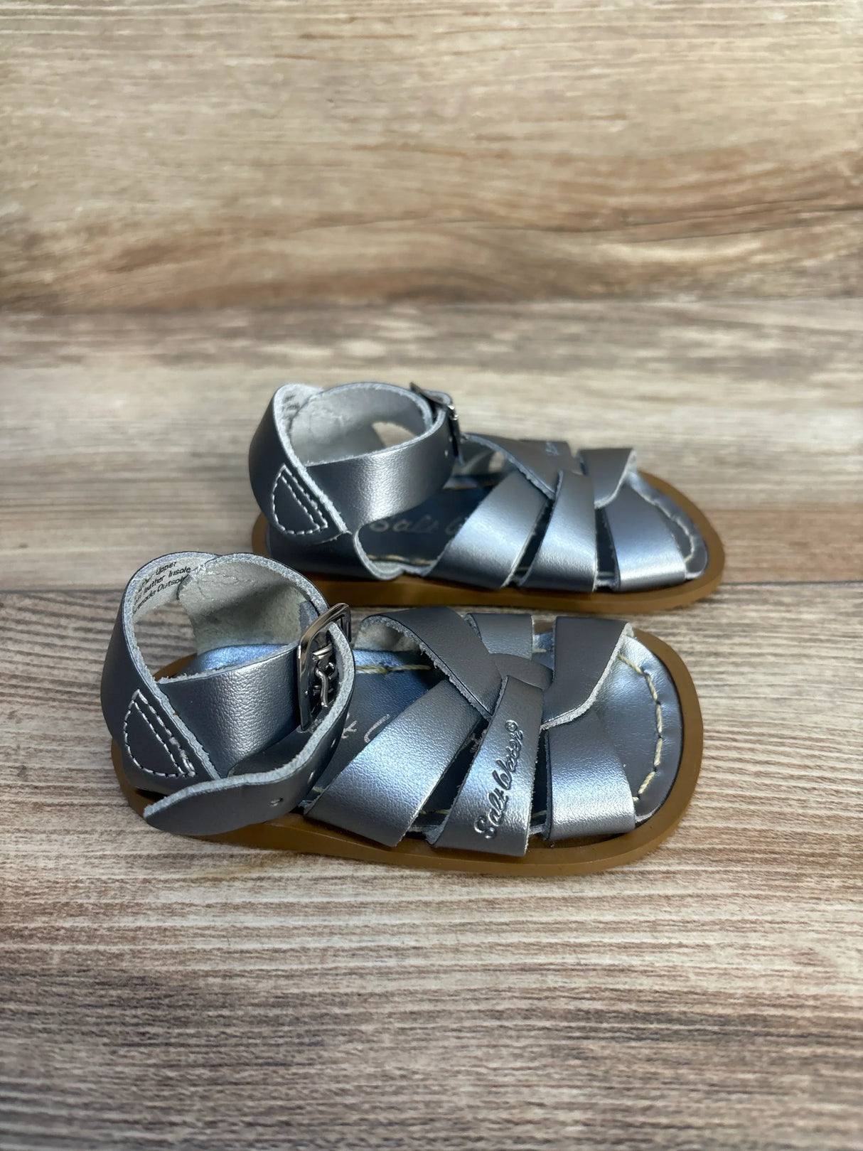 Silver baby sandals with buckled straps on wooden surface, gently used kids footwear.