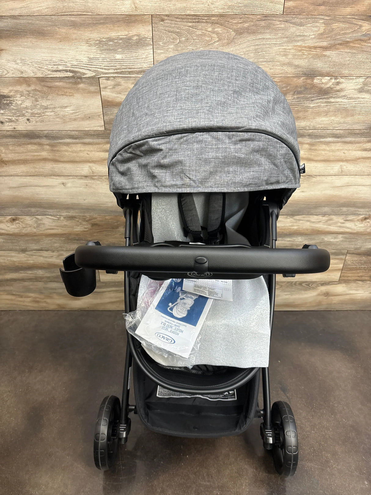 Front view of Graco Modes Nest stroller in Nico showing gray canopy, child tray with cup holder and instruction manual on wood floor