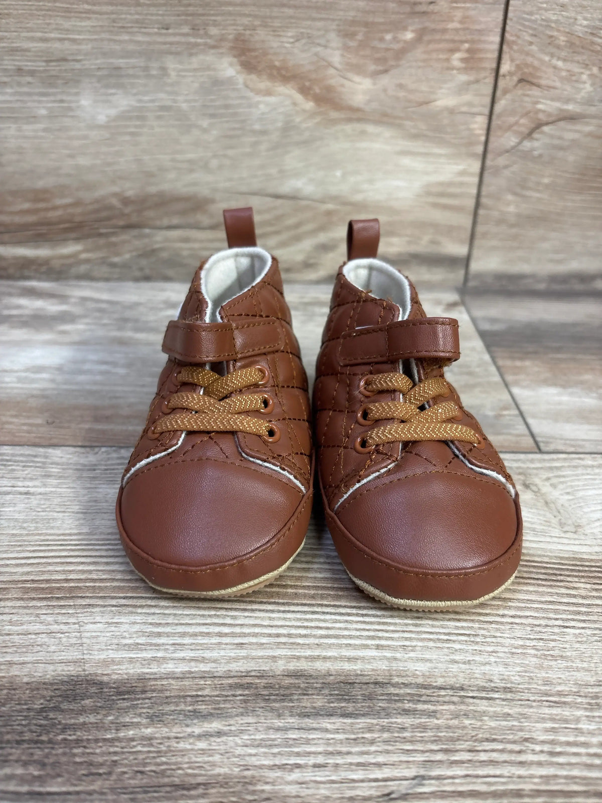 Front view of faux leather crib shoes in brown, size 4C, showcasing fastening straps and laces on a wood-look surface