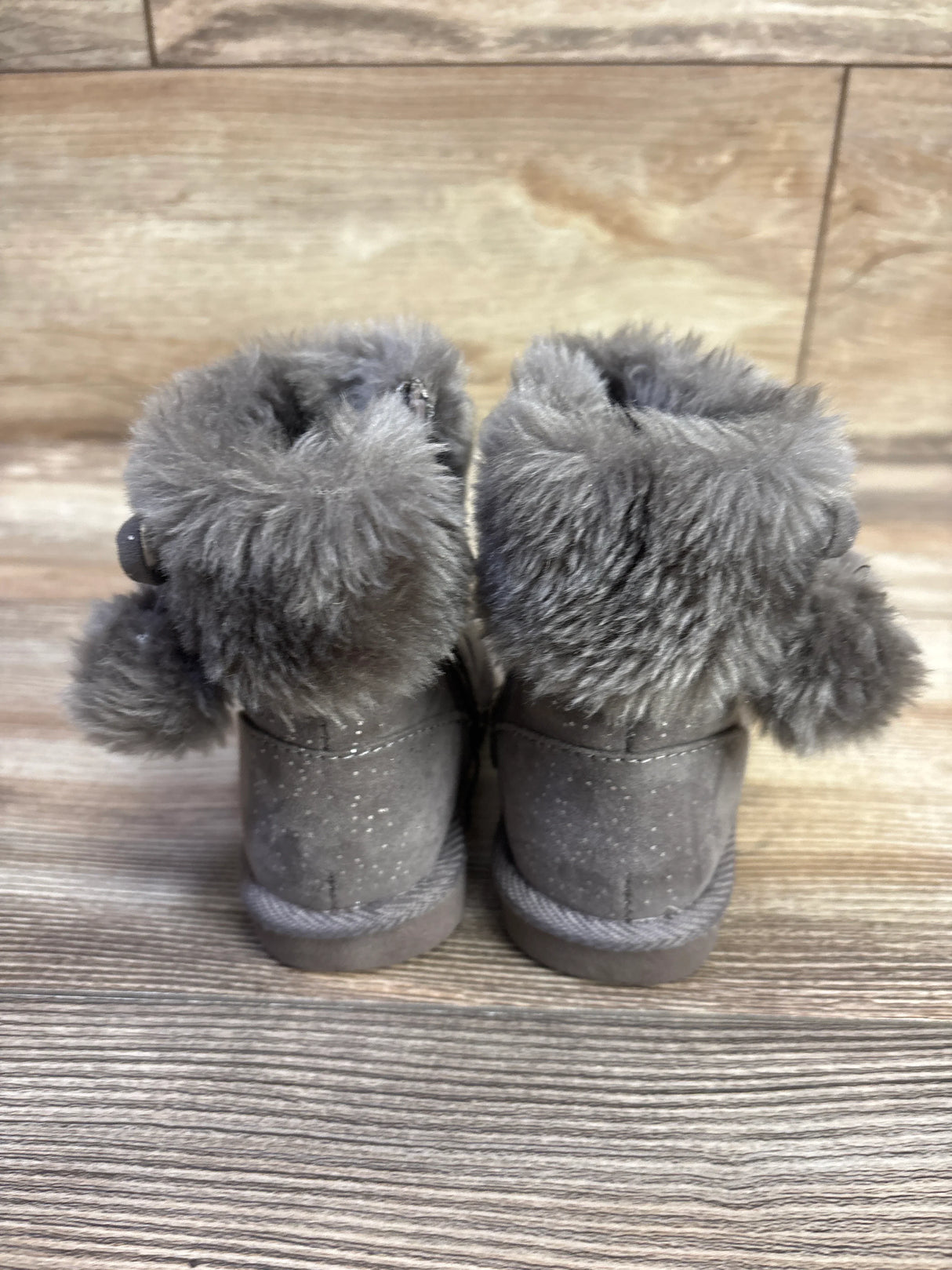 Back view of Jumping Beans Coco Easy Zip toddler boots in grey showing faux fur cuff, pom-poms, and speckled suede on wood background