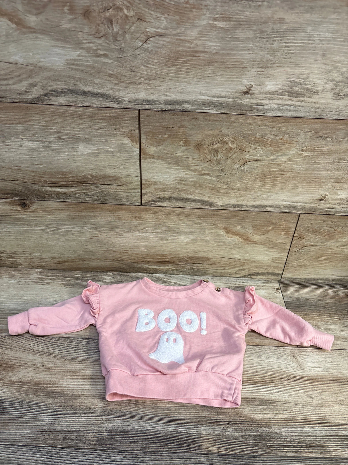 Front flat-lay of pink Jessica Simpson baby sweatshirt with white BOO! and ghost appliqué, ruffled shoulders, snap buttons on wood