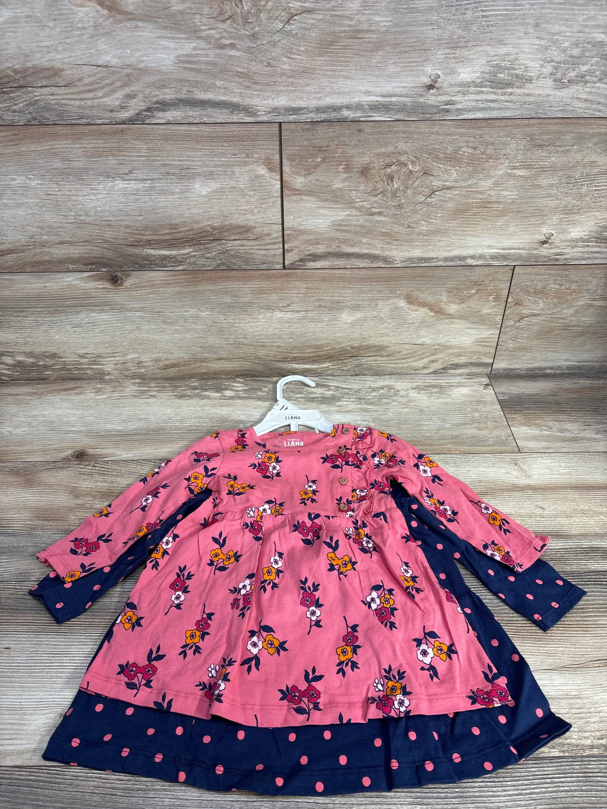 Front view of Laughing Llama dress 2 pack showcasing pink floral and navy polka dot prints layered on hanger on wood plank floor