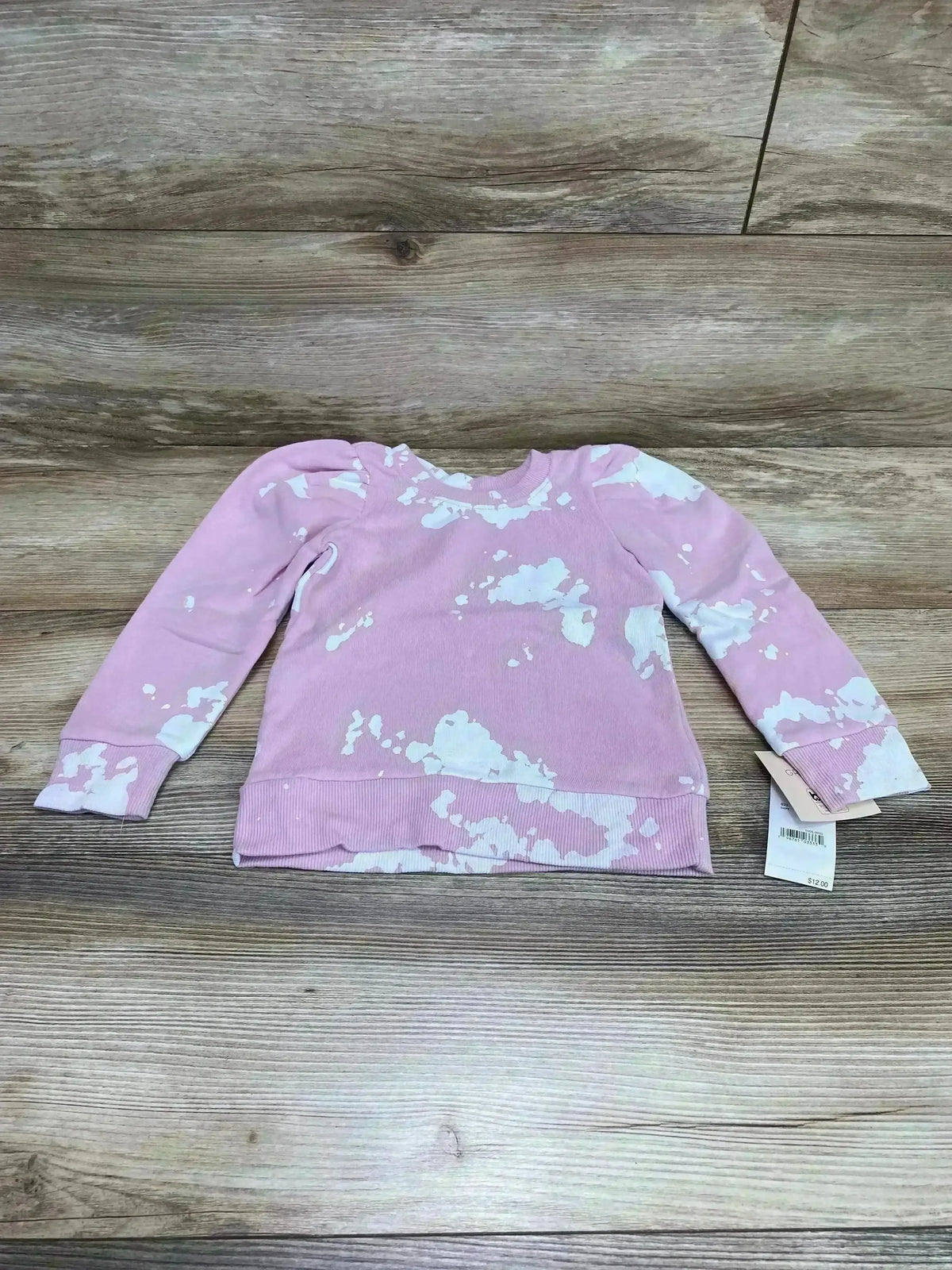 Flat lay of pink Grayson mini puff-sleeve sweatshirt with white splatter pattern and visible 2T tag on wood background