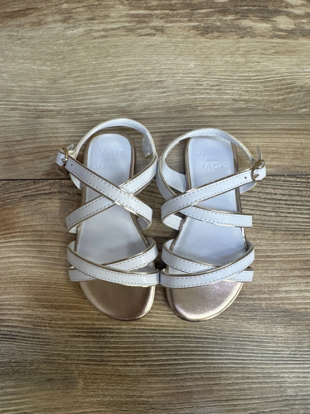 Top view of Janie and Jack white sandal size 7C showing metallic rose gold toe, crisscross straps and ankle buckle on wood