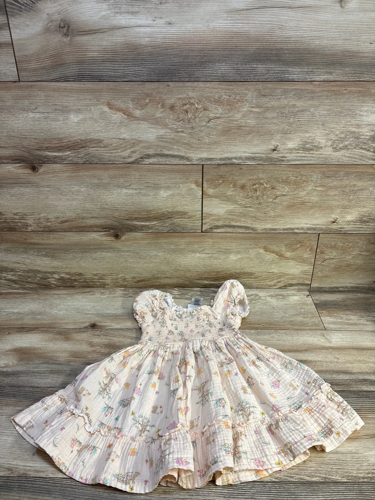Flat lay of Bonnie Jean smocked gauze dress in beige, smocked bodice, flutter sleeves, ruffled hem on wood backdrop