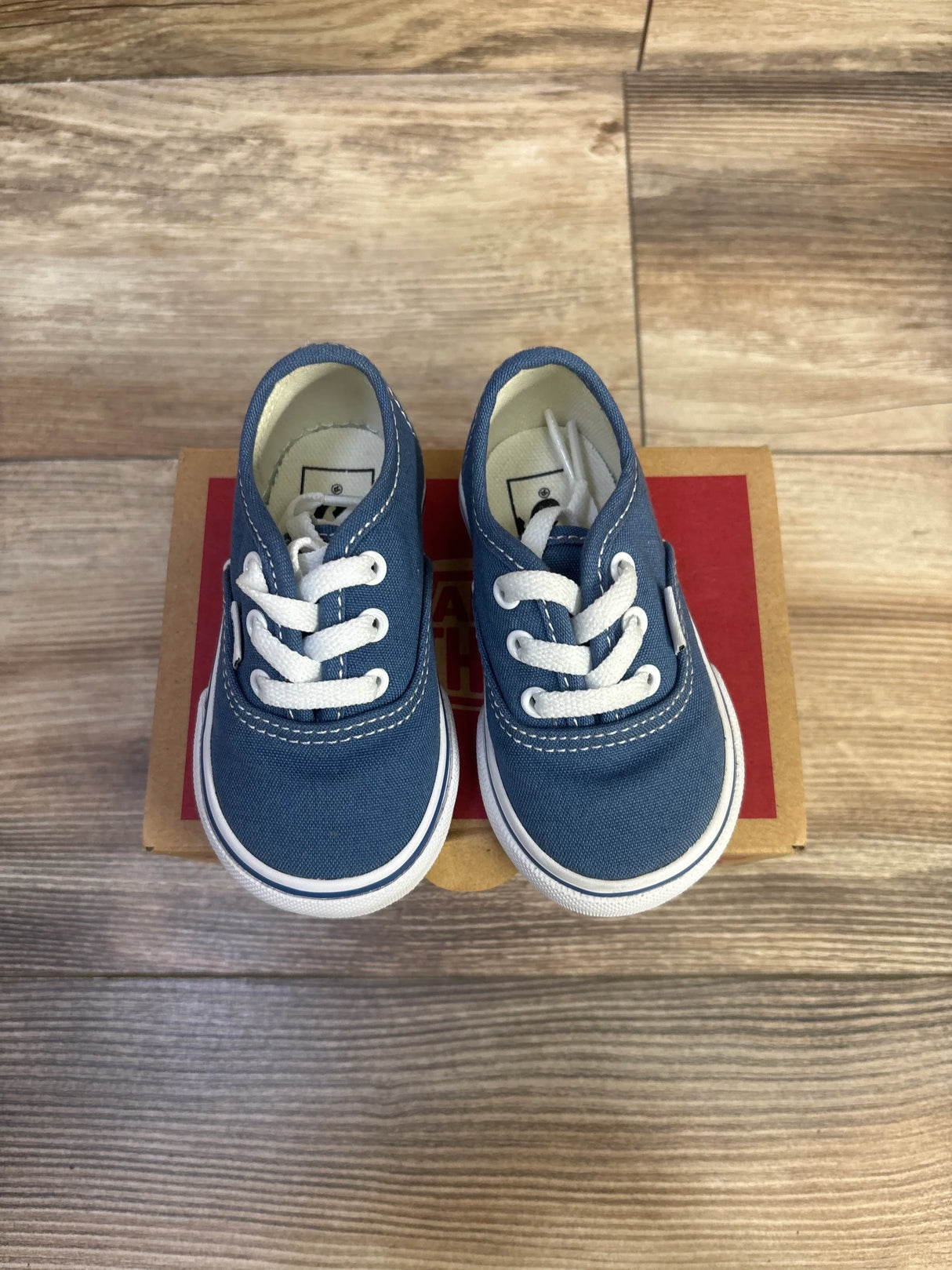Top-down view of navy Vans Authentic low-top toddler sneakers showing white laces and rubber sole on a red box on wood floor