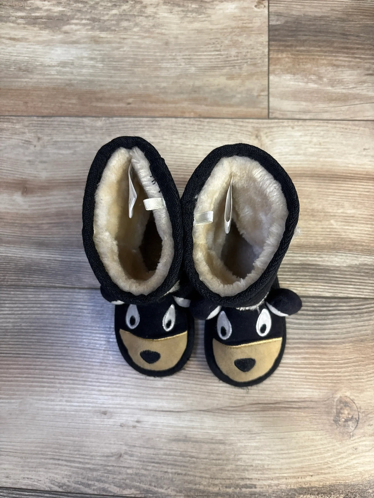 Top-down view of pair of black kids’ bear slipper boots showing plush cream lining, embroidered face and ears on a wood floor