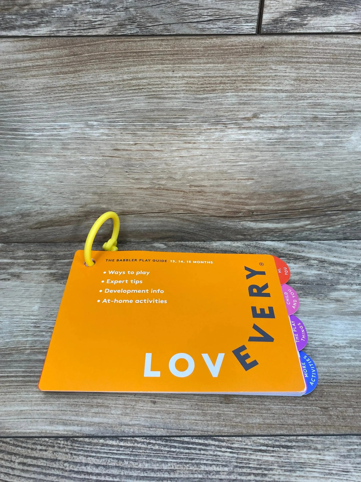 Top-down view of Lovevery The Babbler play guide (orange) showcasing tabbed board pages, yellow plastic ring, and cover text 'Ways to play, Expert tips, Development info, At-home activities' on a wood surface