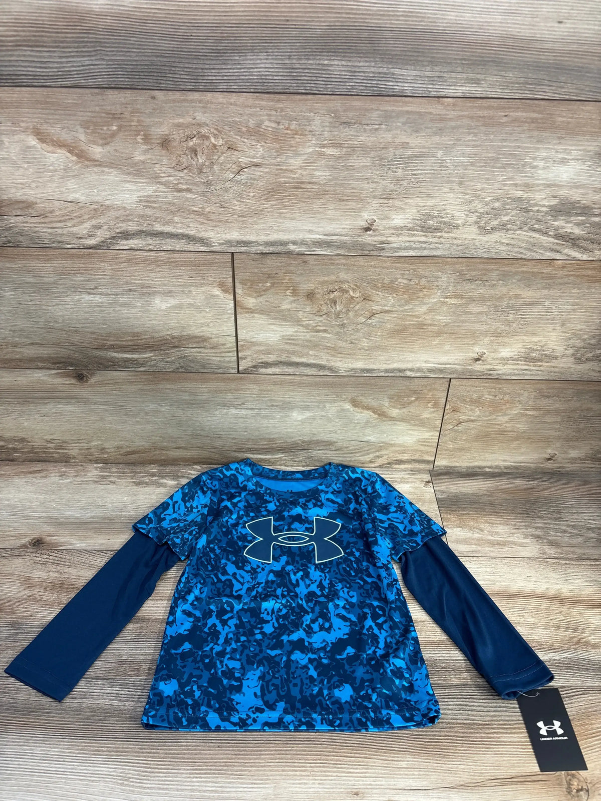 Front flat-lay of Under Armour mock-layered shirt showing blue camo print, navy sleeves, and white logo on wood background