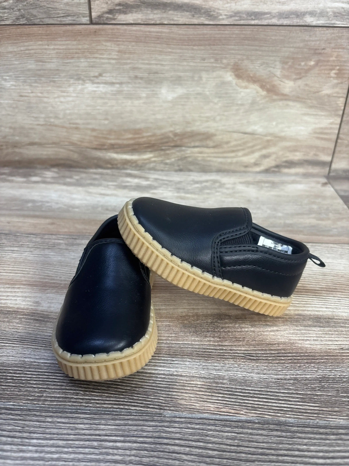 Angled view of OshKosh Victor sneakers showcasing black slip-on design and tan ridged soles on a wood-look surface