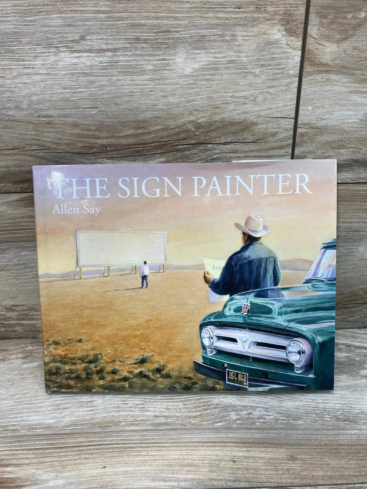 Front cover of The Sign Painter hardcover book showing a man with a hat by a green truck and empty billboard