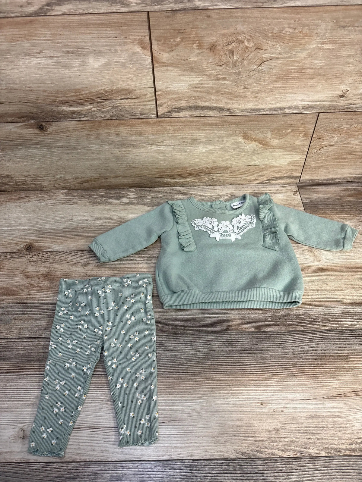 Flat lay of sage green Nicole Miller two-piece sweatshirt set showcasing ruffle shoulders, embroidered chest and floral leggings on wood floor