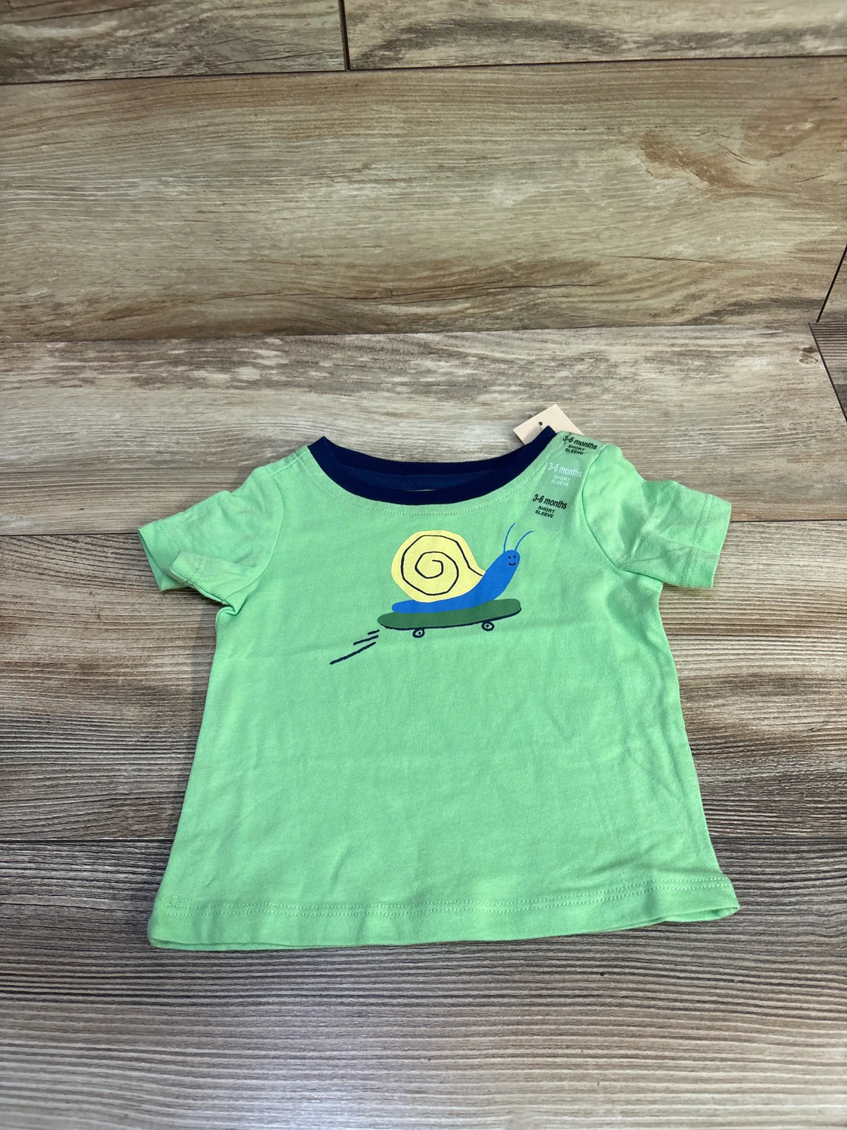 Flat lay of green baby T-shirt (3-6 months) showing skateboarding snail graphic on wood background
