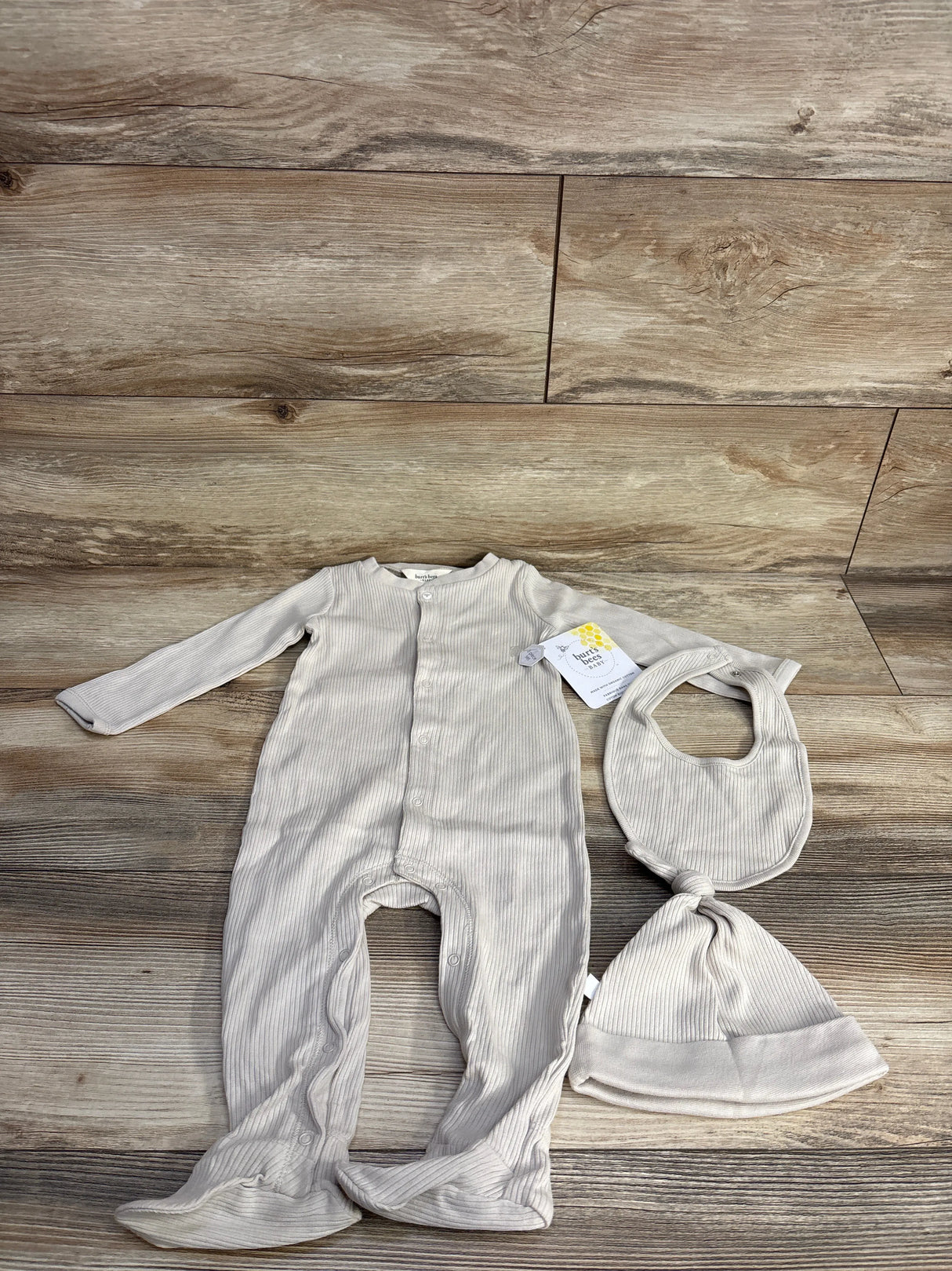 Beige ribbed baby outfit set with footed sleeper, bib, and knotted hat on wooden floor