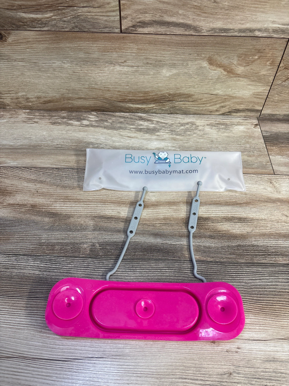 Top-down view of Busy Baby mini mat toy straps in pink showing two gray adjustable straps and three pink suction cups on a wood floor