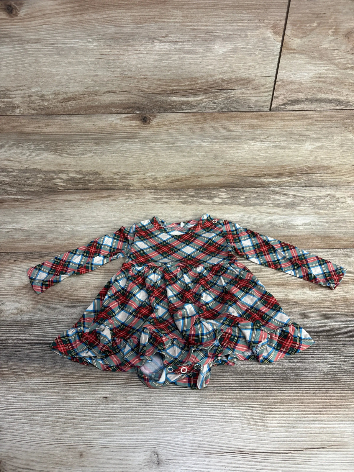 Plaid baby dress with ruffled skirt, long sleeves, and snap closures on wood floor