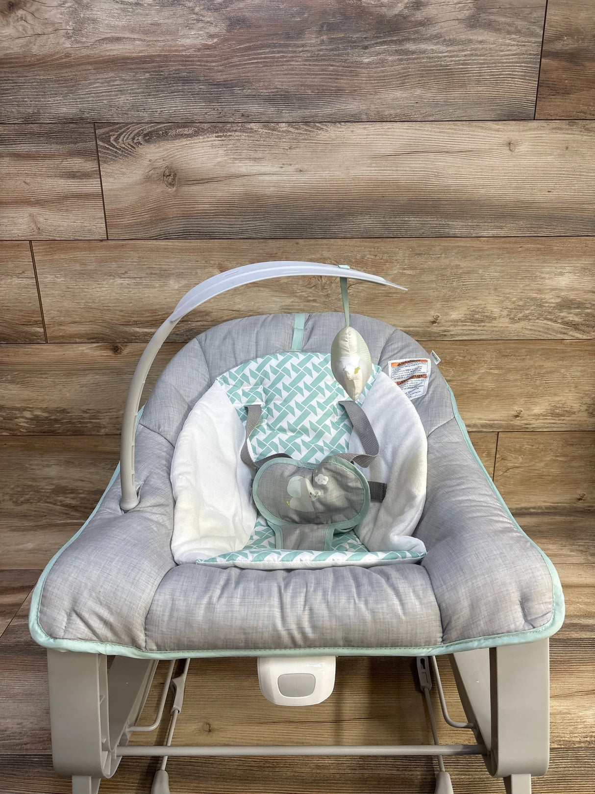 Front view of Ingenuity 3-in-1 baby bouncer with gray padded seat mint geometric insert overhead toy on wood background